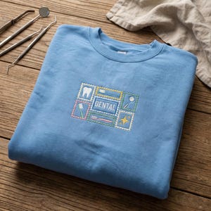 May include: A pale blue sweatshirt with a dental-themed embroidered patch. The patch features a tooth, toothbrush, dental tools, and the word "DENTAL" in a rectangular frame. The design is in pastel colours.