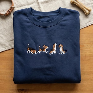May include: A navy blue sweatshirt with an embroidered design of four beagle dogs in various poses. The dogs are brown, black, and white. A dog collar and a bone are visible in the background.