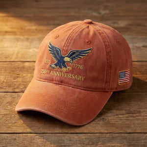 May include: A rust-colored baseball cap featuring an embroidered blue and gold eagle graphic. The text reads EST. 1776 and 250TH ANNIVERSARY. A small embroidered American flag patch is located on the side of the cap, which has a structured crown and brim.