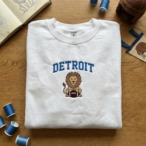 Könnte beinhalten: Weißes Sweatshirt mit Rundhalsausschnitt und dem Wort "DETROIT" in Blau über einem gestickten Löwen, der einen Football hält. Der Löwe ist braun und beige. Das Sweatshirt liegt auf einer Holzoberfläche mit blauen Garnrollen.