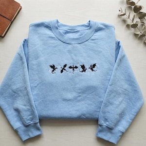 Puede incluir: Sudadera azul claro con cuello redondo. Presenta un dise&ntilde;o bordado de cinco dragones negros con alas extendidas, intercalados con peque&ntilde;os detalles blancos en forma de estrella. La sudadera es de material suave.
