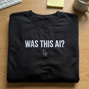 Könnte beinhalten: Schwarzes Sweatshirt mit Rundhalsausschnitt und dem gestickten Text "WAS THIS AI?" in Weiß, mit einer roten und weißen Cursor-Grafik. Das Sweatshirt liegt gefaltet auf einer Holzoberfläche.