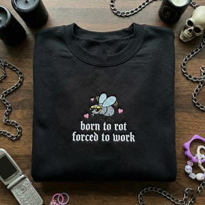 May include: Black crewneck sweatshirt with an embroidered fly wearing sunglasses and hearts. The text "born to rot forced to work" is below the fly. The sweatshirt is surrounded by chains, candles, and a skull.