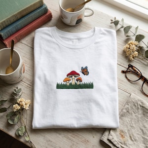 May include: White t-shirt with an embroidered design of mushrooms and a butterfly. The mushrooms have red, orange, and tan caps with green grass. A blue and orange butterfly is near the mushrooms.