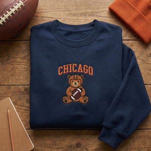 May include: Navy blue sweatshirt with the word "CHICAGO" in orange lettering above a cartoon bear wearing a football helmet and holding a football. An orange beanie and a football are also visible.