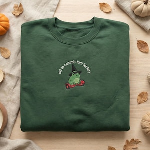 May include: Dark green sweatshirt featuring an embroidered frog in a witch hat on a red hoverboard, with the text "off to commit tom foolery". The sweatshirt is displayed on a wooden surface with autumn decorations, including leaves and pumpkins.