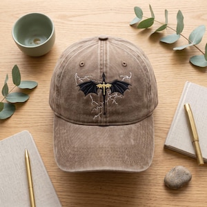 May include: A brown baseball cap featuring an embroidered design of a black dragon with gold wings centered on the front. The dragon is surrounded by white embroidered lightning bolt patterns. The cap has a structured crown and a curved brim.
