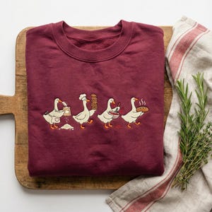 May include: A burgundy sweatshirt with an embroidered design of four geese baking. The geese are shown carrying flour, baked goods, a mixing bowl, and a loaf of bread. The sweatshirt is folded on a wooden chopping board.