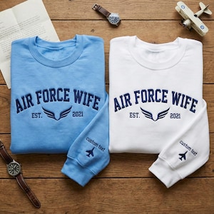 May include: Two crewneck sweatshirts, one light blue and one white, with "AIR FORCE WIFE" embroidered in navy blue. Each has "EST. 2021" and a wing design. Sleeves read "custom text" with a small airplane.