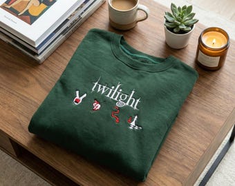 May include: A dark green sweatshirt with the word "twilight" embroidered in white, along with four small embroidered symbols. The sweatshirt is folded on a wooden table, next to a cup of coffee, a succulent, a candle, and a stack of books.