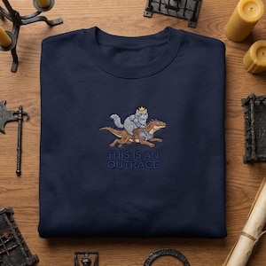 May include: Navy blue sweatshirt with an embroidered design. The design features a gray cat wearing a crown riding a brown dinosaur. The text "THIS IS AN OUTRAGE" is embroidered below the image.