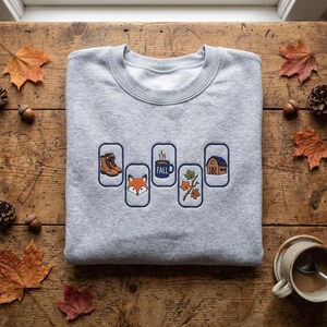 May include: A light grey crewneck sweatshirt featuring five vertical rectangular embroidered patches. The designs include brown hiking boots, a fox face, a blue mug labelled FALL, a branch with orange and green leaves, and a brown barn.