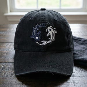 May include: A black baseball cap featuring an embroidered design of two koi fish arranged in a circular yin and yang pattern. One fish is dark blue with white accents, and the other is light gray with dark gray details, centered on the front panel.
