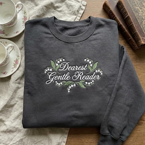 May include: A charcoal grey sweatshirt featuring the embroidered phrase "Dearest Gentle Reader" within a floral wreath. The design incorporates green leaves and white flowers. The sweatshirt has a crew neck.