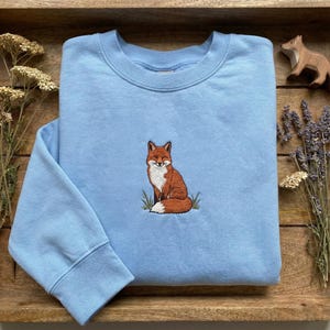 Peut inclure: Un sweat-shirt bleu clair avec une broderie de renard. Le renard est représenté en marron, blanc et orange, assis dans de l'herbe verte. Le sweat-shirt a un col rond et des manches longues, et est fait d'une matière douce.