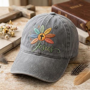 May include: Grey baseball cap featuring an embroidered floral design and the word "Jesus" in green script. The floral design has a cross at the centre and colourful petals. The cap is made of a soft, worn fabric.