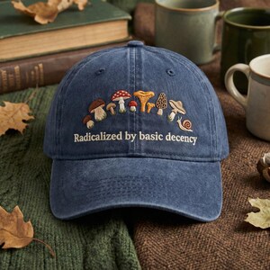 May include: A navy blue baseball cap featuring an embroidered design of different mushrooms and a snail. The text "Radicalized by basic decency" is embroidered below the mushroom design. The cap is on a green and brown surface.