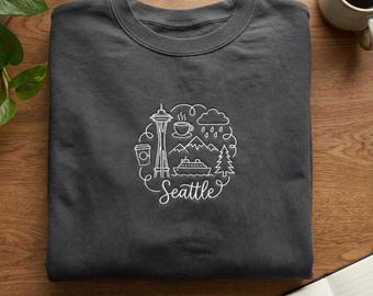 May include: Dark grey sweatshirt featuring a white graphic of Seattle landmarks, including the Space Needle, mountains, coffee cup, and a ferry. The word "Seattle" is written in cursive. The sweatshirt is folded on a wooden surface.