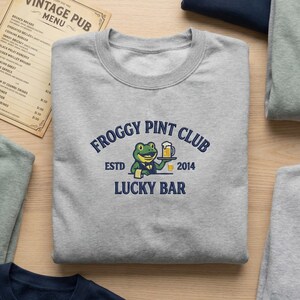 May include: A light gray sweatshirt with the text "FROGGY PINT CLUB" and a cartoon frog holding a beer. The sweatshirt also has the text "ESTD 2014 LUCKY BAR". A vintage pub menu is visible in the background.
