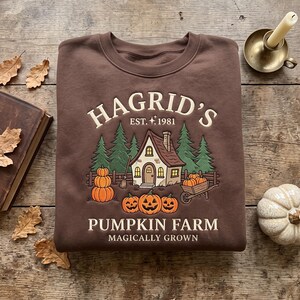May include: A brown crewneck sweatshirt featuring a graphic print of a cottage, pine trees, and pumpkins. The text reads HAGRID'S EST. 1981 PUMPKIN FARM MAGICALLY GROWN. The design includes a wheelbarrow, a broom, and jack-o'-lanterns in orange and green.