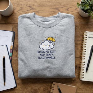 May include: A grey crew neck sweatshirt with an embroidered design. The design shows a smiling cloud under a yellow umbrella, with the navy text: "DOING MY BEST AND THAT'S QUESTIONABLE".