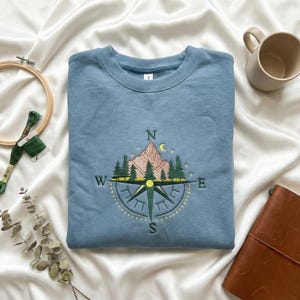 May include: A blue sweatshirt with an embroidered compass design featuring a mountain, trees, and a crescent moon. The compass points to North, South, East, and West. The sweatshirt is folded on a white satin surface.