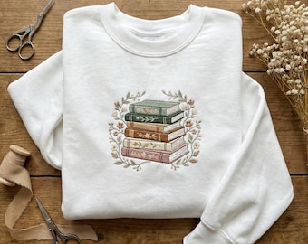 May include: White sweatshirt featuring an embroidered design of a stack of books in shades of green, brown, and pink, surrounded by a floral wreath. The design is centered on the chest. The sweatshirt is made of a soft material.