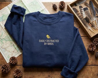May include: Navy blue sweatshirt with the embroidered text "EASILY DISTRACTED BY BIRDS" and a yellow bird graphic. The sweatshirt is folded on a wooden surface with a map, pinecones, and a tray of feathers.