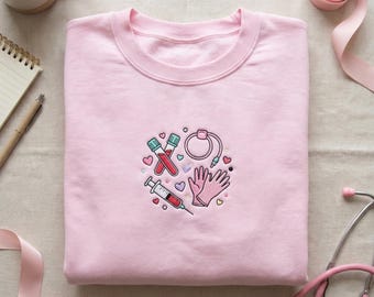 May include: A pale pink sweatshirt featuring an embroidered medical design. The design includes test tubes with blood, a syringe, medical gloves, and a medical device, all surrounded by hearts. The sweatshirt is folded neatly on a light-coloured surface.