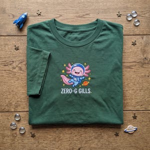 May include: A dark green t-shirt featuring a graphic of a pink axolotl wearing a blue astronaut suit floating in space. The design includes yellow stars and a small orange planet. Below the graphic, the white text reads ZERO-G GILLS.