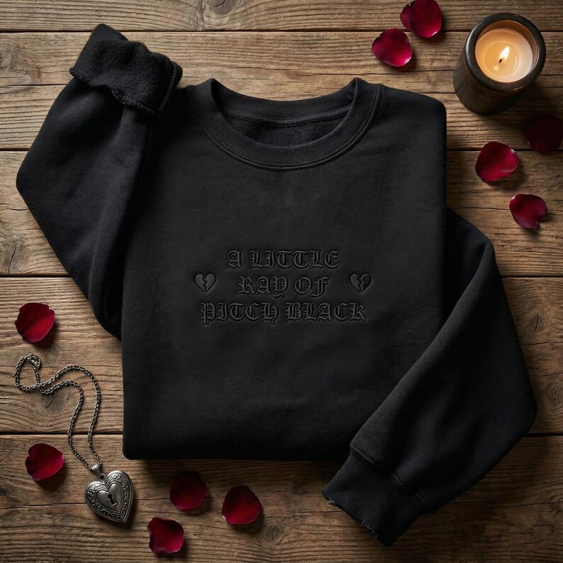 May include: Black crewneck sweatshirt with the text "A Little Ray of Pitch Black" in gothic font. The sweatshirt is surrounded by rose petals, a lit candle, and a heart-shaped locket necklace on a wooden surface.