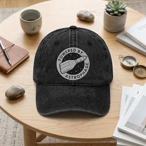 May include: Black baseball cap with a distressed finish. The cap features a circular embroidered design with the words "POWERED BY ASTROPHAGE" and a rocket ship illustration. The phrase "HAIL MARY" is also included in the design.