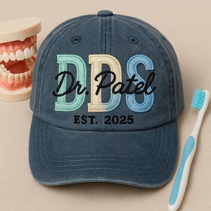 May include: A navy blue baseball cap with the embroidered text "DDS Dr. Patel EST. 2025" in teal, beige, and blue. A model of teeth and a blue and white toothbrush are next to the cap. The cap is a dental-themed accessory.