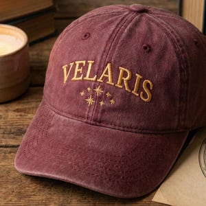 May include: A maroon baseball cap with the word "VELARIS" embroidered in gold thread. Below the word are embroidered stars. The cap is sitting on a wooden surface next to a lit candle and a celestial map.