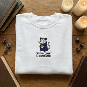 May include: White crew neck sweatshirt with an embroidered opossum in a blue cloak, holding a gold emblem. The text "OFF TO COMMIT SHENANIGANS" is below. The image is set against a background of candles and dice.