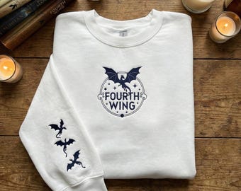 May include: White crewneck sweatshirt with a navy blue dragon graphic and the text "FOURTH WING". The left sleeve features three navy blue dragon silhouettes. The sweatshirt is surrounded by candles and books.