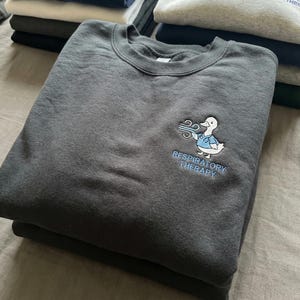 May include: Dark gray sweatshirt with a crew neck. The sweatshirt features an embroidered design of a duck wearing a medical coat with the words "RESPIRATORY THERAPY" in blue below the duck. Several other folded sweatshirts are in the background.