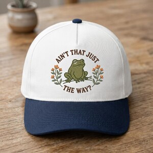 May include: A baseball cap with a white crown and a navy blue brim. The cap features an embroidered design of a green frog and the text "AIN'T THAT JUST THE WAY?" in a circular arrangement.