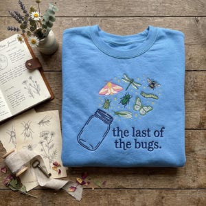 Könnte beinhalten: Ein hellblaues Sweatshirt mit Rundhalsausschnitt und einer Stickerei, die ein Glasgefäß zeigt, aus dem verschiedene bunte Insekten wie ein Falter, eine Libelle, eine Biene und Käfer fliegen, mit dem darunter gestickten Text "the last of the bugs.".