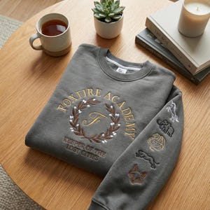 May include: A gray sweatshirt with the text "FOXFIRE ACADEMY" in gold embroidery, encircled by a wreath. The sleeve features embroidered animal designs. A cup of tea, a succulent, a candle, and books are on a wooden table.
