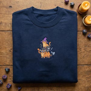 May include: A navy blue sweatshirt with an embroidered design of three cats in fantasy costumes. The cats are dressed as a wizard, a warrior, and a barbarian. The sweatshirt is made of a soft material and is navy blue.