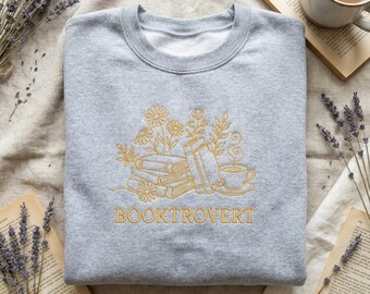 May include: A light gray sweatshirt with a gold embroidered design featuring books, flowers, and a teacup. The word "BOOKTROVERT" is embroidered below the design. The sweatshirt is displayed on a neutral-toned surface with dried lavender and open books.