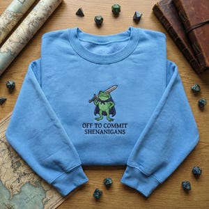 May include: A pale blue sweatshirt with a round neck. It has an embroidered design of a cartoon frog with a sword and cape. The text "OFF TO COMMIT SHENANIGANS" is embroidered below the frog.