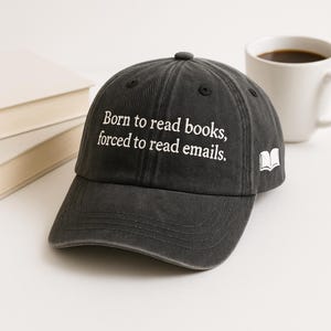 Peut inclure: Casquette de baseball gris foncé avec le texte "Born to read books, forced to read emails" en blanc. Une petite icône de livre blanc est sur le côté. Une tasse à café blanche et une pile de livres sont en arrière-plan.