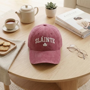 May include: A dusty rose baseball cap with the word "SLÁINTE" embroidered in white, along with a small shamrock. The cap is sitting on a wooden table, alongside a cup of tea, a teapot, and a stack of books.