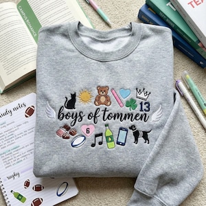 May include: A gray sweatshirt embroidered with the words "boys of tommen" and various colorful icons, including a cat, sun, teddy bear, heart, and dog. The number 13 is also featured. The sweatshirt is surrounded by books and stationery.