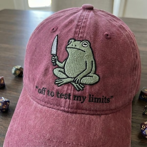 May include: A maroon baseball cap with an embroidered design of a green frog holding a knife. The frog is embroidered with a detailed texture. The text "off to test my limits" is embroidered below the frog.