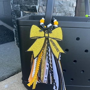 May include: A black tote bag with a decorative keychain featuring a large gold glitter bow, black and white ribbons, and yellow beads. The bow has the word WARRIORS printed on it, and the ribbons include text that reads CHEER.