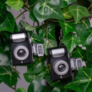 May include: A pair of black camera earrings with silver accents, hanging against a backdrop of green ivy. The earrings feature detailed miniature camera designs, including lenses and flashes, perfect for photography enthusiasts.