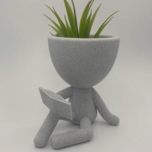 May include: A grey ceramic planter shaped like a person sitting with a book in their lap. The planter has a textured surface and a small green plant growing out of the top.